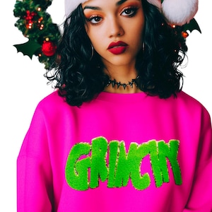 May include: A bright pink sweatshirt with the word "GRINCH" in green, textured lettering. The person in the image is wearing a white Santa hat with a fluffy pom-pom and a black choker. A Christmas tree is in the background.