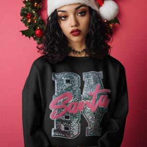 May include: Black sweatshirt with the text "BA Santa BY" in a sequined design. The word "Santa" is in pink, and the letters "BA" and "BY" are in silver sequins. A white Santa hat with a red pom-pom is worn.