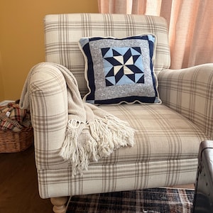 Peut inclure: Un fauteuil à carreaux beiges avec un coussin orné d'un motif d'éclat d'étoile bleu et blanc. Une couverture à franges est drapée sur le bras. Un tapis à motifs et un panier sont visibles en arrière-plan.