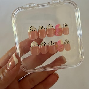 May include: A set of ten press-on nails in a clear plastic storage case. The nails feature a nude base with black polka dots on the tips. Two of the nails are accented with a raised, three-dimensional pink floral design and a small gold bead.