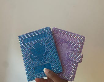May include: Two passport covers, one blue and one lavender, are decorated with sparkling rhinestones. The blue cover features the United States of America seal. The word "PASSPORT" is visible on both covers. The covers are held by a hand.