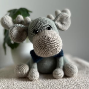 May include: A crocheted gray and blue moose plush toy with black eyes. The moose is sitting on a light brown fuzzy surface.