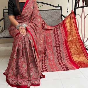 May include: A red and gold saree with a traditional Indian print. The saree is made of a soft, flowing fabric and has a beautiful drape.