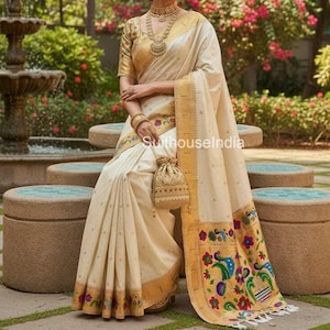 Puede incluir: Una mujer con un sari color crema con detalles dorados y bordados intrincados. El sari presenta un borde detallado con diseños florales y de pavo real coloridos. También lleva joyas de oro, incluyendo un collar, aretes y pulseras. Un pequeño bolso cuelga de su mano.