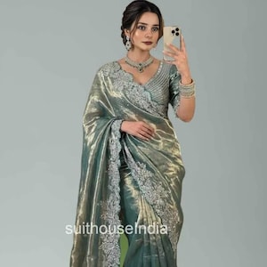 Könnte beinhalten: Ein schimmernder, salbeigrüner Sari mit aufwendiger silberner Stickerei an den Rändern und am Ausschnitt. Der Stoff hat einen metallischen Glanz, der ein luxuriöses Aussehen erzeugt. Der Sari ist elegant drapiert und zeigt sein fließendes Design. Der Text "suithouseindia" ist sichtbar.
