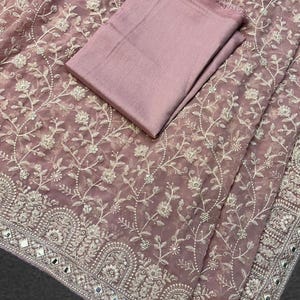 May include: A dusty rose-colored saree with intricate white floral embroidery. The saree features a detailed border with mirrored accents. A matching fabric piece is folded and placed on top of the saree.