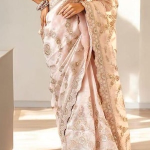 Pode incluir: Um sari rosa claro com bordados e enfeites dourados intrincados. O sari apresenta uma borda detalhada e uma blusa combinando com um padrão listrado. O tecido parece transparente e fluido, criando um visual elegante e formal.