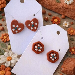 May include: Terracotta-colored clay earrings in heart and hexagon shapes, each with white and teal accents. The earrings are displayed on white tags, surrounded by decorative flowers and clay elements.