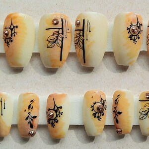 May include: Set of 20 false nails with a light orange and white ombre design. The nails feature black floral and geometric designs with gold accents.