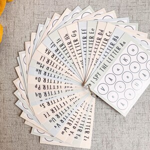 May include: A set of alphabet flashcards fanned out, featuring the text "I Spy The Letter" and individual letters. The cards are in pastel colors and are attached with a metal ring. The background is a neutral gray fabric.
