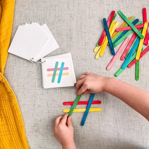 Könnte beinhalten: Ein Kind verwendet bunte Holzstäbchen, um das Hashtag-Symbol zu erstellen. Die Stäbchen sind auf einer grauen Oberfläche angeordnet. Im Hintergrund ist ein Kartenset mit verschiedenen Symbolen zu sehen.