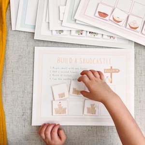 May include: A child's hand interacting with a "Build a Sandcastle" educational activity set. The set includes illustrated cards and a guide with instructions. The background features additional activity sheets and a yellow fabric.