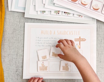 Puede incluir: Una mano infantil interactuando con un juego educativo "Construye un castillo de arena". El juego incluye tarjetas ilustradas y una guía con instrucciones. El fondo presenta hojas de actividades adicionales y una tela amarilla.