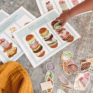 May include: Educational food-themed activity set featuring illustrated cards for building a waffle tower and a burger. Includes cut-out food pieces like waffles, fruit, and burger ingredients. A child's hand is reaching for a food piece.