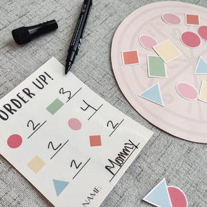 Pu&ograve; includere: Un cartoncino bianco con le parole "Order Up!" e una griglia di numeri e forme. Il cartoncino &egrave; su una superficie grigia con un cerchio rosa con varie forme e colori. Il testo "Mommy" &egrave; scritto sul cartoncino.