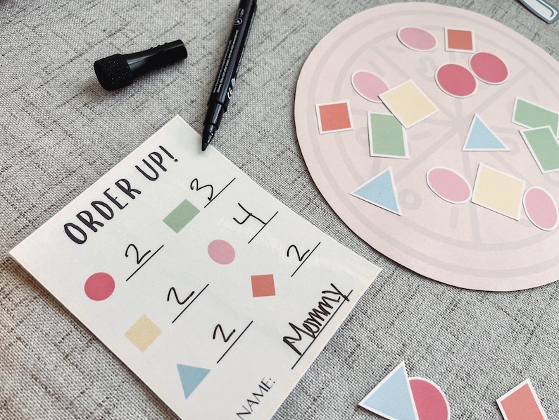 Pu&ograve; includere: Un cartoncino bianco con le parole "Order Up!" e una griglia di numeri e forme. Il cartoncino &egrave; su una superficie grigia con un cerchio rosa con varie forme e colori. Il testo "Mommy" &egrave; scritto sul cartoncino.