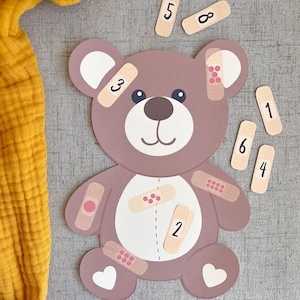 Può includere: Un puzzle a forma di orsetto marrone con bende e numeri da 1 a 8. L'orso ha una faccia, orecchie e piedi a forma di cuore bianchi. Le bende sono beige con accenti rosa e rossi. I numeri sono neri.