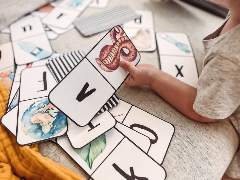 Pu&ograve; includere: Una mano di bambino tiene una carta bianca con una lettera V nera e un'immagine di un serpente marrone. La carta fa parte di un set di carte flash dell'alfabeto con varie illustrazioni.
