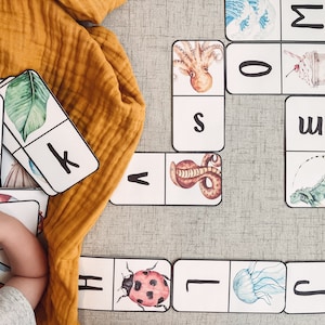 Puede incluir: Un juego de dominó de colores con imágenes de animales y letras. Los dominós están dispuestos sobre una superficie gris. Los dominós están hechos de cartulina gruesa y tienen un acabado brillante.