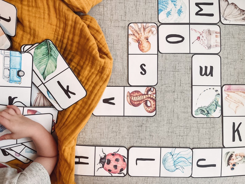 Pu&ograve; includere: Un set di domino colorati con immagini di animali e lettere. I domino sono disposti su una superficie grigia. I domino sono fatti di cartoncino spesso e hanno una finitura lucida.