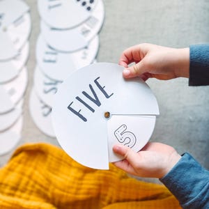 May include: A white circular card with the word "FIVE" written in black. The card is divided into sections, with the number "5" written in black on one section. The card is being held by a child's hands.