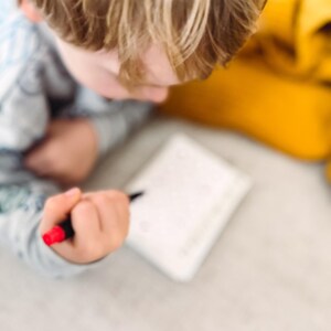 May include: A child's hand holding a black pen with a red tip, writing on a white notepad. The child's blond hair is visible, along with a yellow item in the background. A red marker is on the surface.