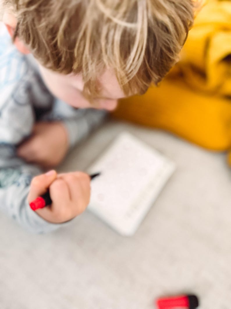 May include: A child's hand holding a black pen with a red tip, writing on a white notepad. The child's blond hair is visible, along with a yellow item in the background. A red marker is on the surface.