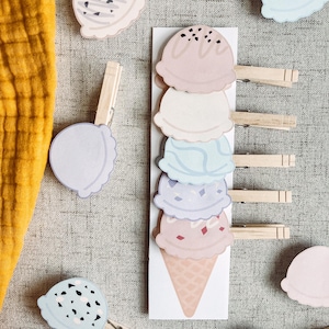 Può includere: Un set di mollette di legno con disegni colorati di coni gelato. I coni gelato sono impilati su una carta bianca con un disegno di cono a cialde. Le mollette sono un modo divertente e fantasioso per aggiungere un tocco di estate alla tua decorazione domestica.
