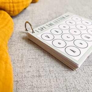 May include: A small, spiral-bound activity book with the text "I SPY THE LETTER A" printed on the cover. The pages feature a grid of circles with letters inside. The book is held together by a silver metal ring. A yellow fabric is in the background.