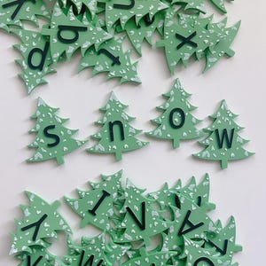 May include: Green Christmas tree-shaped wooden letters with black letters and white accents. The letters spell "SNOW" and are scattered among other letters. The letters are likely for holiday decor or crafting.