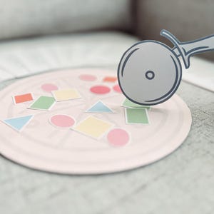 Pu&ograve; includere: Un tabellone da gioco a forma di pizza rosa con forme colorate e un taglia pizza in cima.