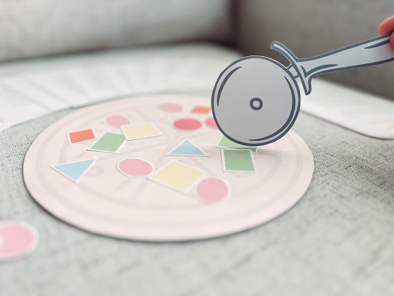 Pu&ograve; includere: Un tabellone da gioco a forma di pizza rosa con forme colorate e un taglia pizza in cima.