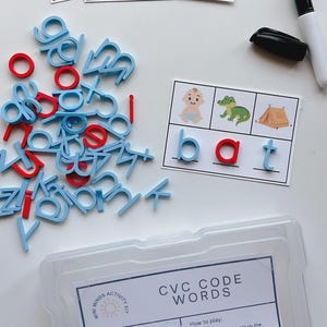 May include: A children's educational activity kit with CVC code words. The kit includes blue and red lowercase letters, cards with pictures, and a dry-erase marker. The clear plastic container has the text "CVC CODE WORDS".