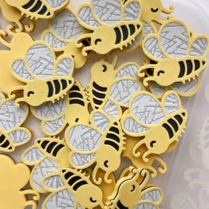 May include: A collection of yellow bee-shaped embellishments with black stripes and gray wing details. The bee-themed items are in a clear plastic container. The bees have a cartoon-like design.