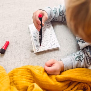 May include: A child's hand using a red marker to circle a letter on a small spiral-bound notebook. The notebook reads "I SPY THE LETTERS". A red marker cap sits next to the notebook. A yellow blanket is in the foreground.