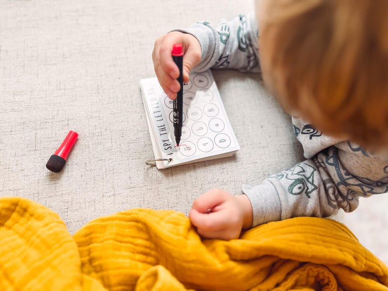 May include: A child's hand using a red marker to circle a letter on a small spiral-bound notebook. The notebook reads "I SPY THE LETTERS". A red marker cap sits next to the notebook. A yellow blanket is in the foreground.