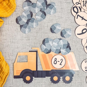 Op de afbeelding: Een gele speelgoed-kipper met het nummer 8-2 aan de zijkant, die grijze stenen vervoert. De vrachtwagen staat op een grijs, getextureerd oppervlak. Er liggen extra grijze stenen rond de vrachtwagen.