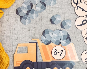 Puede incluir: Un camión de volteo de juguete amarillo con el número 8-2 en el lateral, transportando piedras grises. El camión está sobre una superficie gris texturizada. Hay piedras grises adicionales esparcidas alrededor del camión.