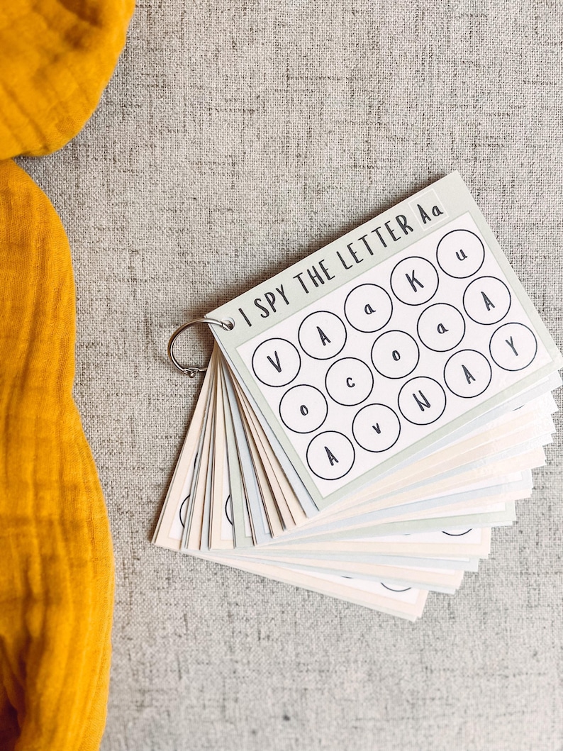 May include: A children's educational activity set with the text "I SPY THE LETTER Aa" on the cover. The pages are bound by a metal ring and feature circles with letters. The background is a light gray fabric, with a yellow cloth in the upper left corner.