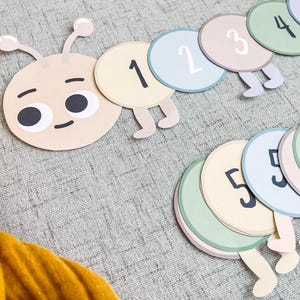 May include: A colorful felt caterpillar with a friendly face and numbers 1 through 5 on its segments. The caterpillar is made of felt and has a light brown head with two large eyes. The segments are in various shades of green, blue, and pink.