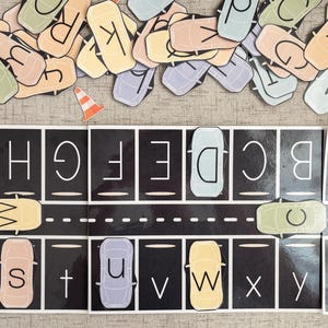 May include: A black and white alphabet learning mat with a road design. The letters A through Z are on individual cars, which are placed in the parking spaces on the mat. The cars are in various colors, including yellow, blue, green, and pink.