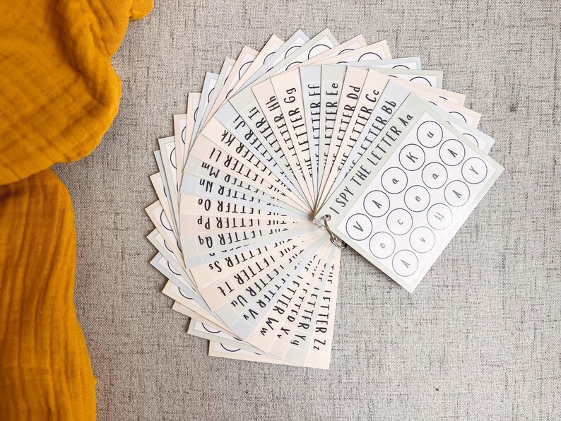 May include: A set of alphabet flashcards fanned out, featuring the text "I Spy The Letter" and individual letters. The cards are in pastel colors and are attached with a metal ring. The background is a neutral gray fabric.