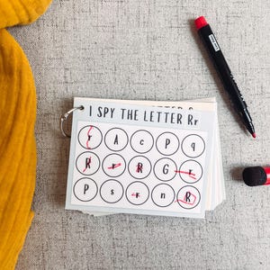 May include: A children's educational game with the text "I SPY THE LETTER Rr". The game includes a stack of cards with circles containing letters, a black marker, and a red eraser. The game is on a gray surface.