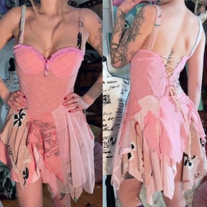 Könnte beinhalten: Ein rosa Kleid mit einem korsettartigen Oberteil und einem mehrschichtigen Rock. Das Oberteil hat einen herzförmigen Ausschnitt und dekorative Verzierungen. Der Rock hat eine Mischung aus gemusterten und einfarbigen rosa Stoffen. Der Rücken des Kleides hat einen Schnürverschluss. Das Kleid hat dünne Träger.