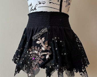 May include: Black mini skirt with a high, elasticated waistband. The skirt features intricate embellishments, including sequins, lace, and a large floral design with a purple gem in the centre. The skirt is displayed on a mannequin.