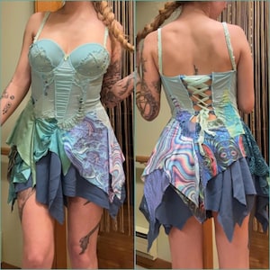 May include: A light blue corset-style dress with a layered skirt. The bodice features delicate embroidery and lace details. The skirt has multiple layers of fabric in shades of blue, green, and patterned designs. The back of the dress has a lace-up closure.