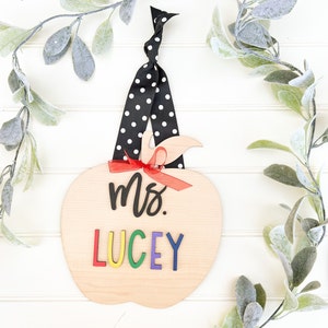 May include: A wooden apple door hanger with a black and white polka dot ribbon. The apple is painted with the text "Mrs. Lucey" in rainbow letters.