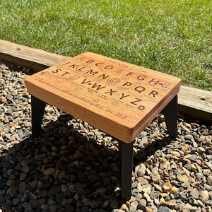 May include: A small wooden stool with four black metal legs and a light brown rectangular top. The top surface features engraved black text including the alphabet from A to Z, the phrase God Says I Am, and the scripture I have loved you with an everlasting love. Jer. 31:3.