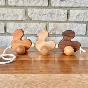May include: A wooden toy pull-along with three duck shapes. The ducks are made of different types of wood, with one being a light brown, one a darker brown, and one a natural wood color. Each duck has a wooden ball for wheels.