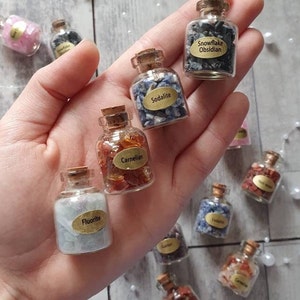 May include: A selection of miniature glass bottles filled with gemstones like Snowflake Obsidian, Sodalite, and Carnelian. Each bottle has a cork stopper and a label with the gemstone's name. The bottles are held in a hand.